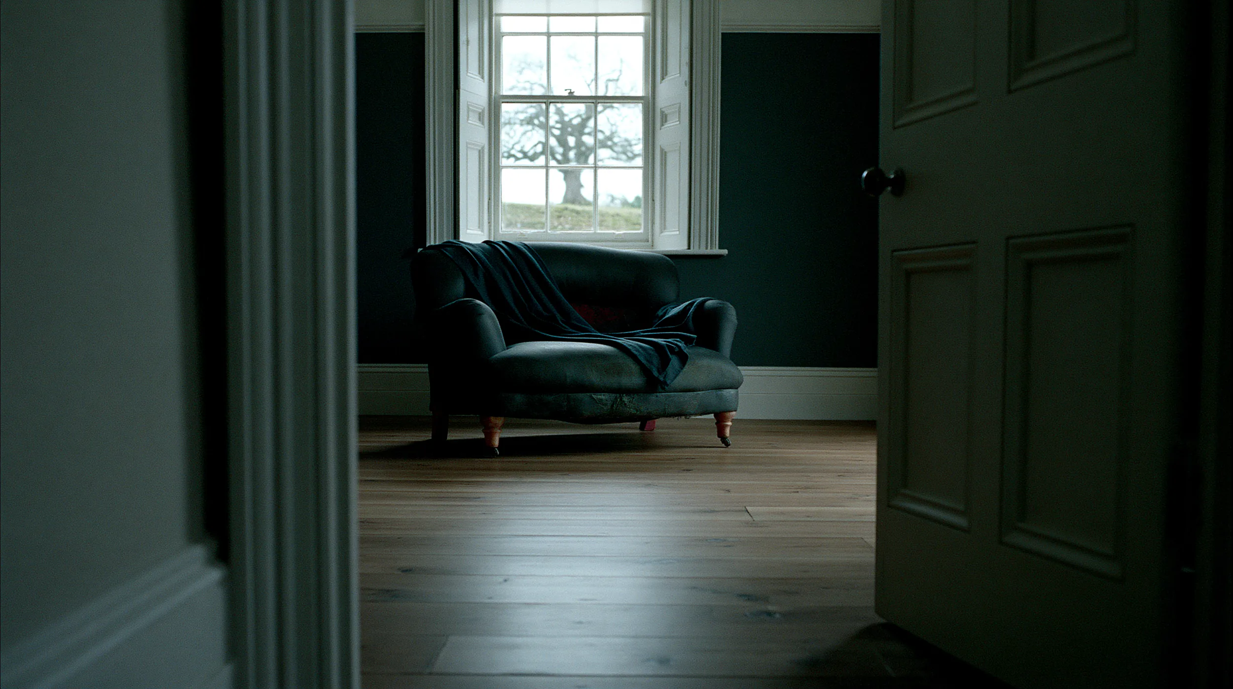 Dark cottagecore living room with teal velvet armchair and sash window overlooking a winter oak tree
