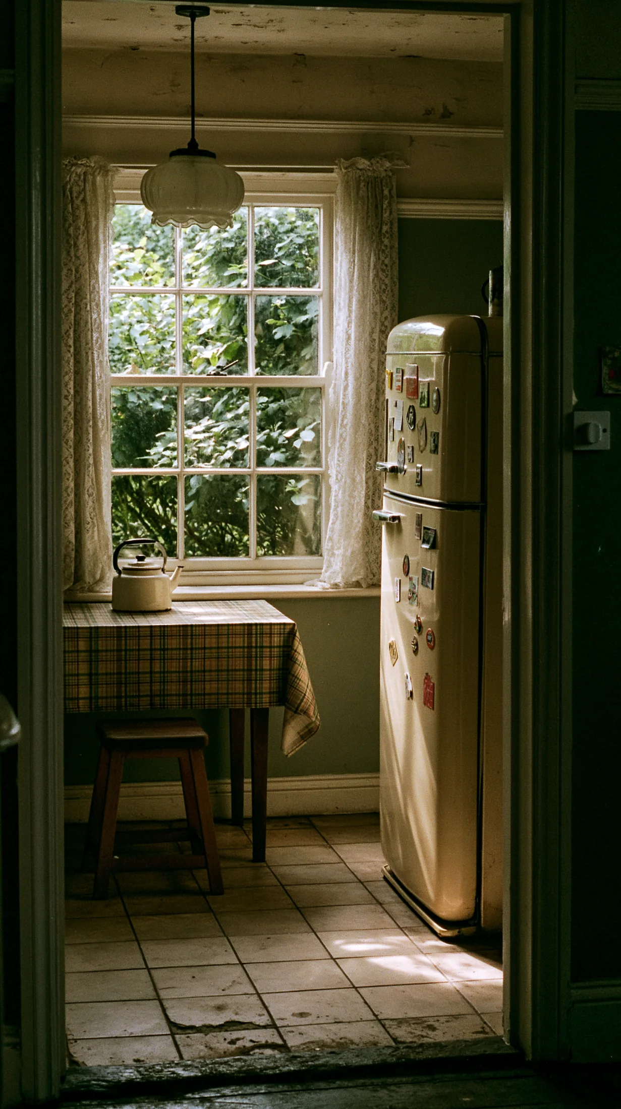 Dark cottagecore kitchen seen through a doorway, with a vintage cream refrigerator covered in magnets, lace curtains, and an enamel kettle on a plaid tablecloth by the window