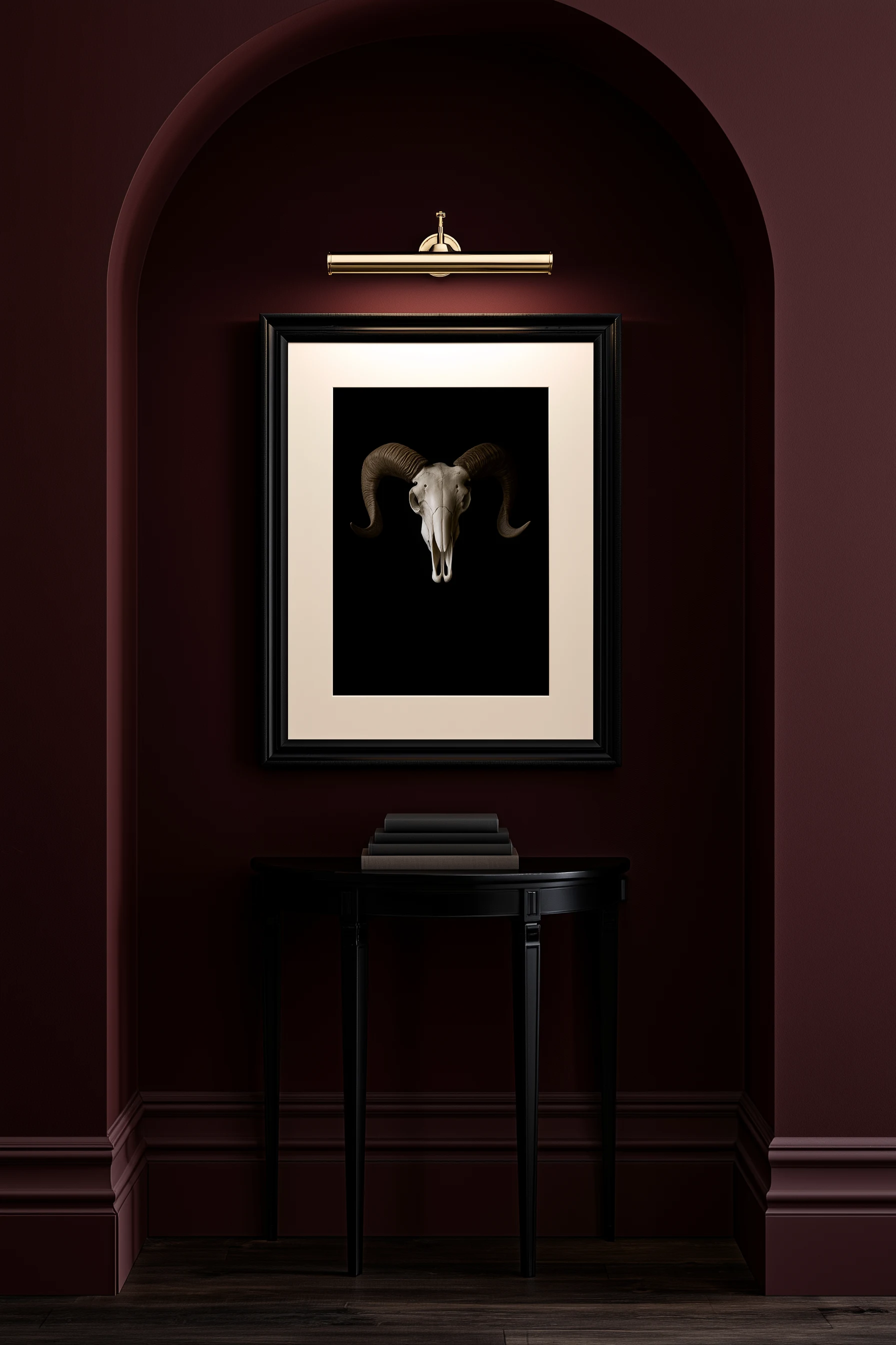 Ram skull photography print in a black frame with a wide cream mat, hung in an arched oxblood niche and lit by a brass picture light, black demilune table with stacked books below