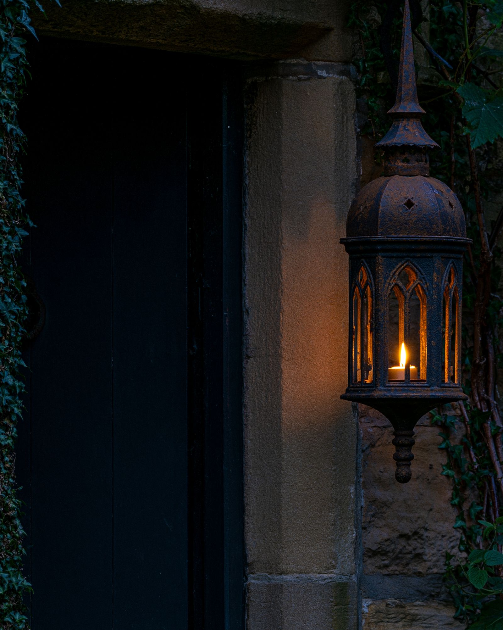 A small cast iron lantern with gothic arched windows hung beside a dark cottage door, one warm light pooling across the stone doorframe at dusk