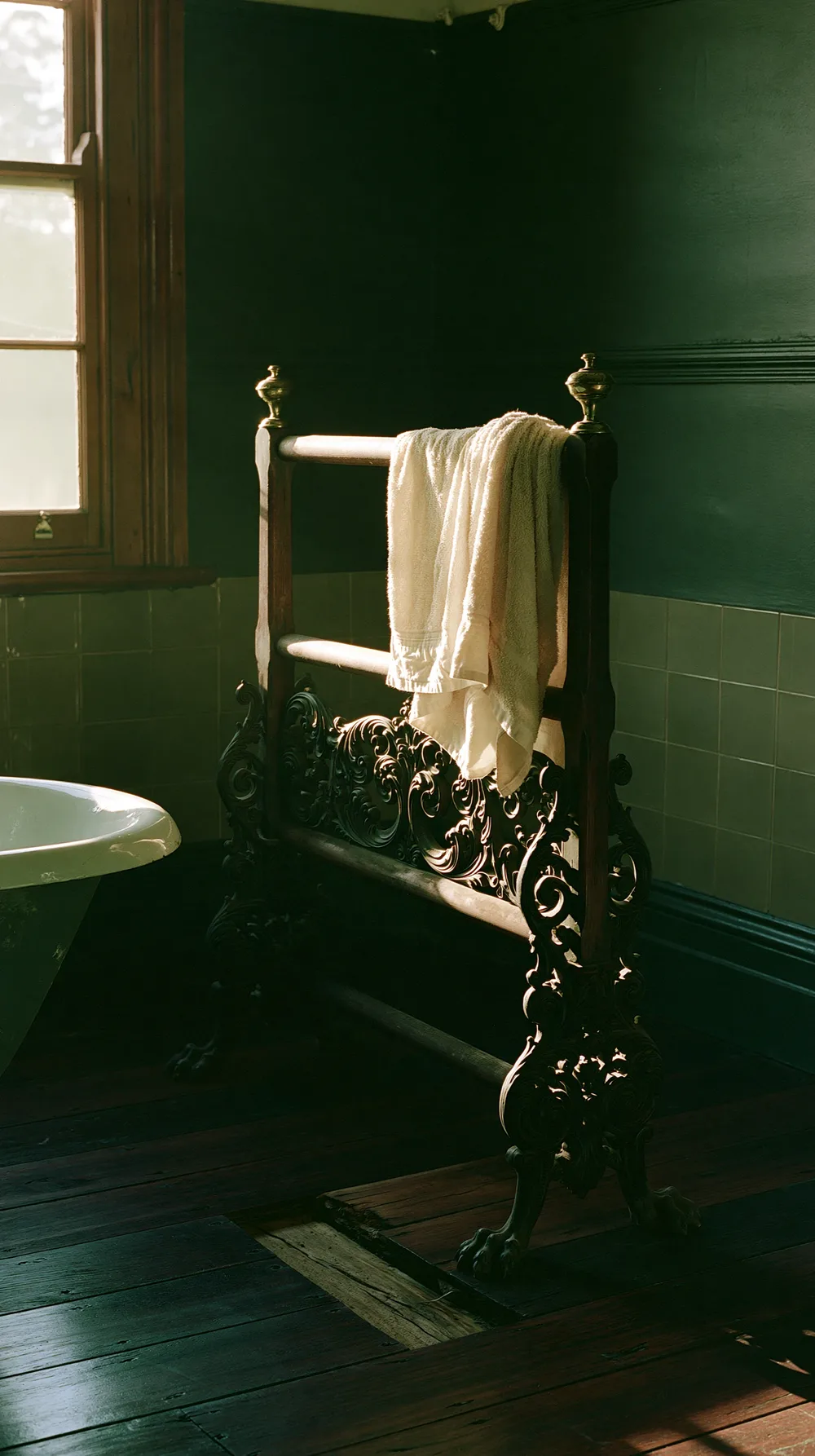 Antique brass towel rack with a linen towel draped over it beside a clawfoot bathtub in a dark green-tiled bathroom lit by afternoon window light