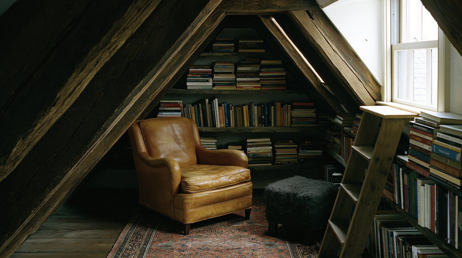 A leather armchair tucked under a sloped attic roofline, surrounded by built-in shelving, a wool pouf, and a leaning library ladder