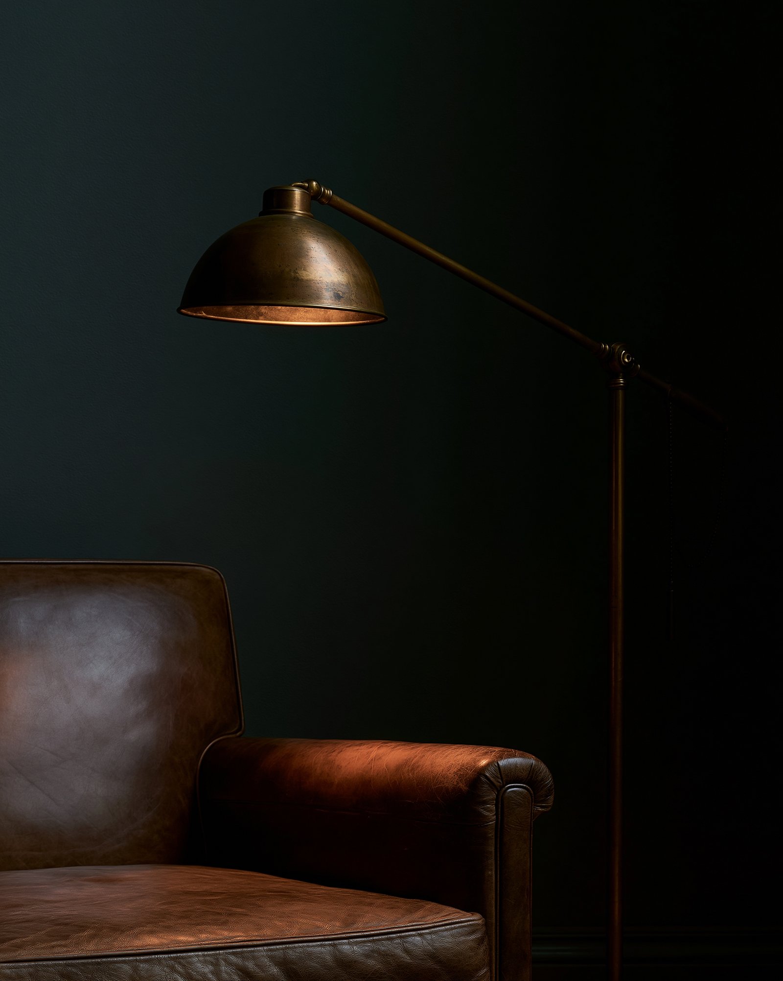 A brass swing-arm floor lamp lit low over a leather armchair's armrest, everything beyond the light falling to true shadow